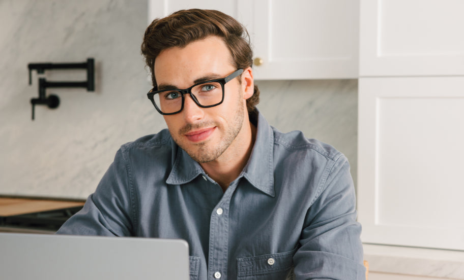 Man wearing glasses and a blue shirt sitting at a table with a laptop in a kitchen.